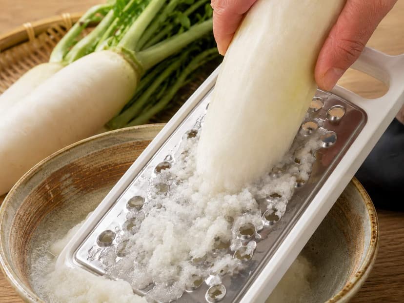 A Japanese daikon grater with a metal grating plate set in a white plastic frame, with grated daikon collecting in the tray below